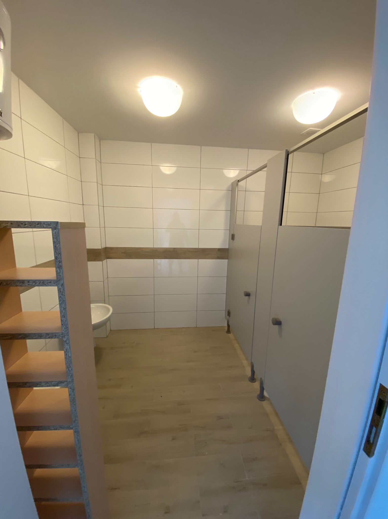 Wnętrze publicznej toalety z kabinami, białe płytki na ścianach, jasna podłoga, widoczny fragment niedokończonego regału z płyty wiórowej.