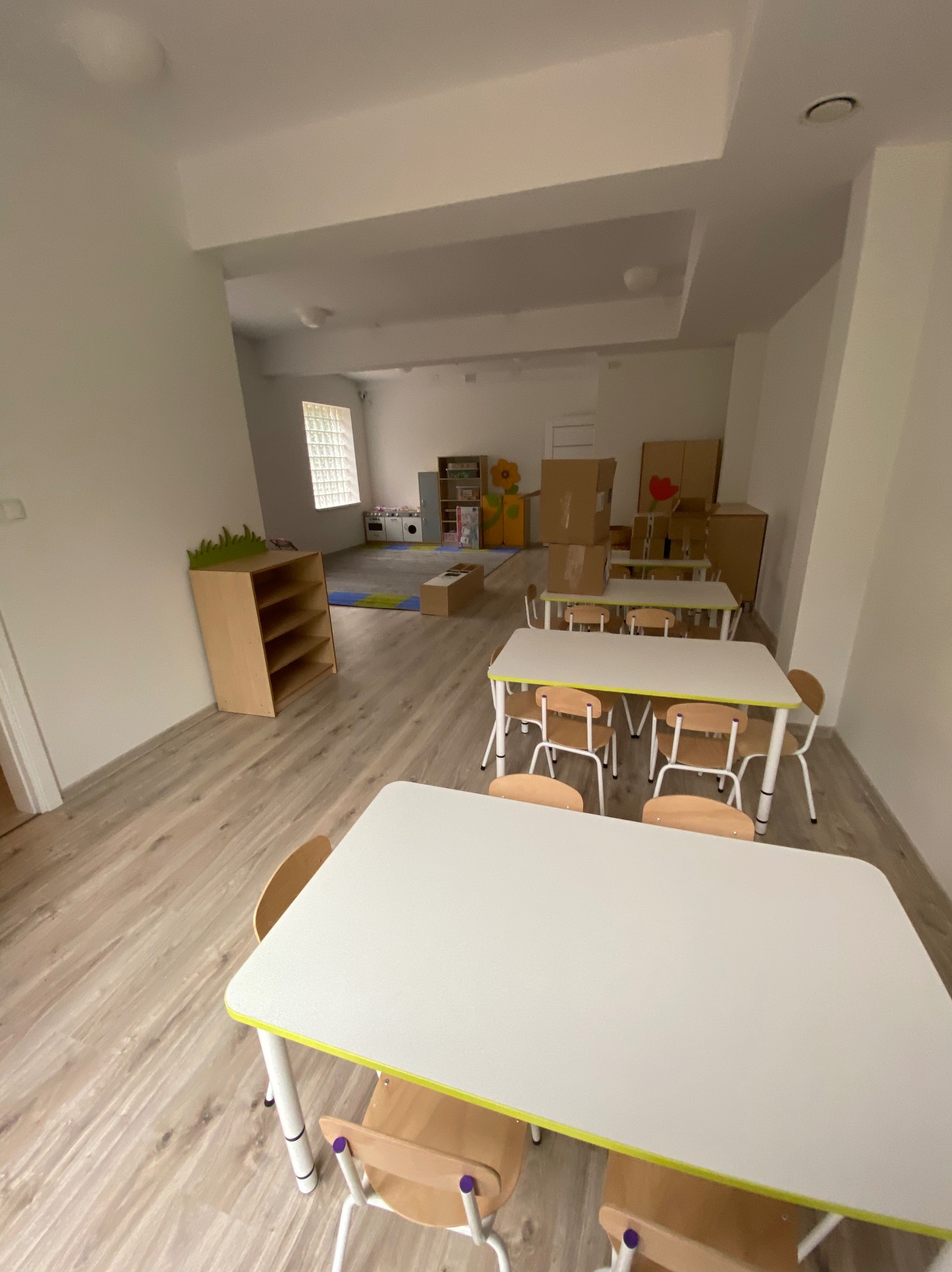 Jasne, przestronne wnętrze sali przedszkolnej z białymi stolikami, drewnianymi krzesłami i laminowaną podłogą imitującą drewno, z widocznym kącikiem zabaw i regałami w tle.