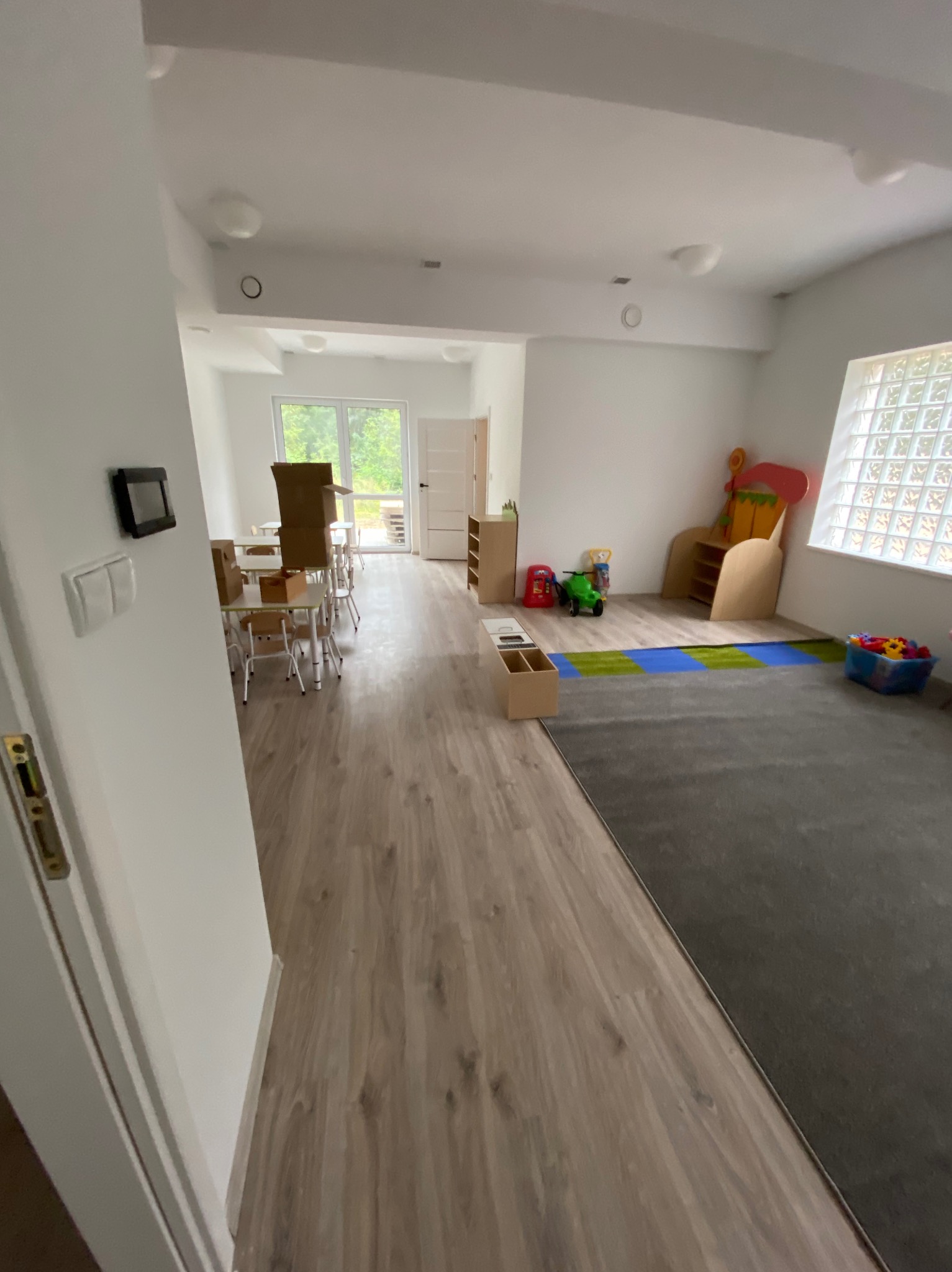Przestronne, nowocześnie wykończone wnętrze sali zabaw z drewnianą podłogą, szarą wykładziną dywanową, białymi ścianami i dużym oknem z luksferami; widoczne zabawki, stoliki z krzesłami i kartonowe...
