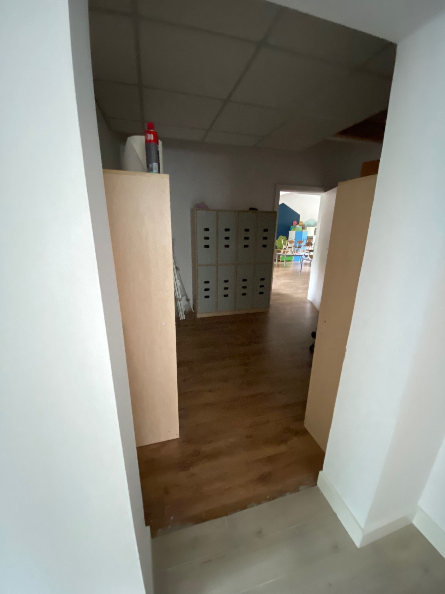 Wnętrze w trakcie remontu: widok na korytarz z brązową podłogą, częściowo zasłonięte szafki z kratkami wentylacyjnymi i widoczne elementy pokoju dziecięcego w oddali.