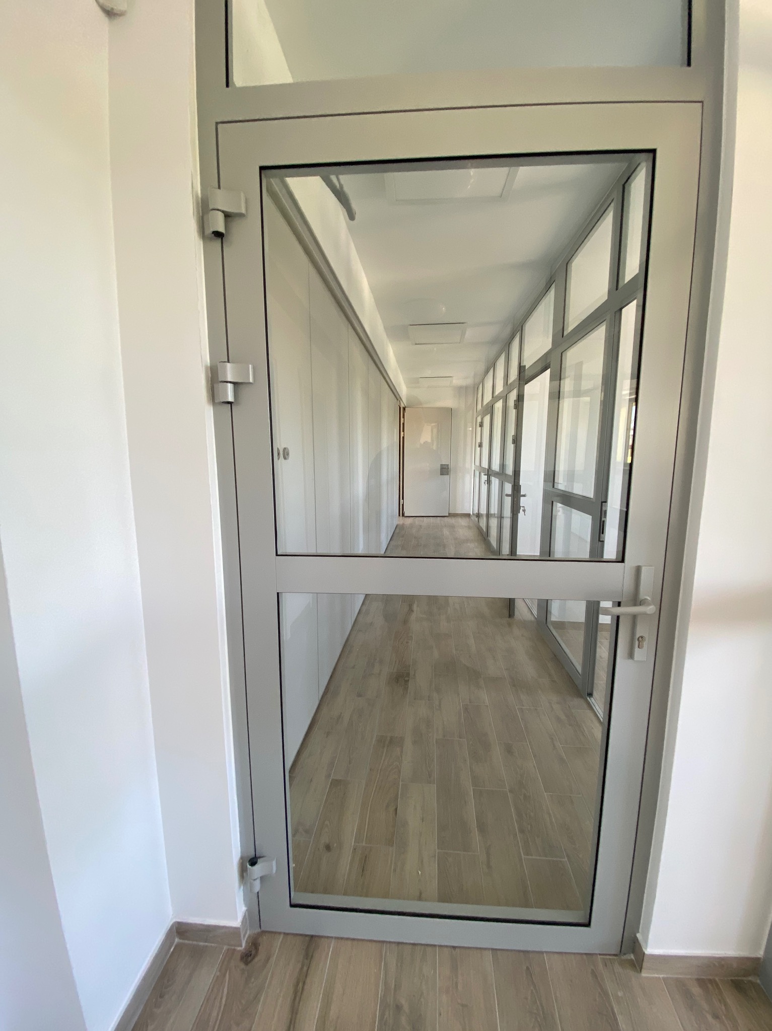 Szklane drzwi w metalowej ramie, widok na długi korytarz z drewnopodobną podłogą i szklanymi ścianami działowymi, białe ściany i sufit.