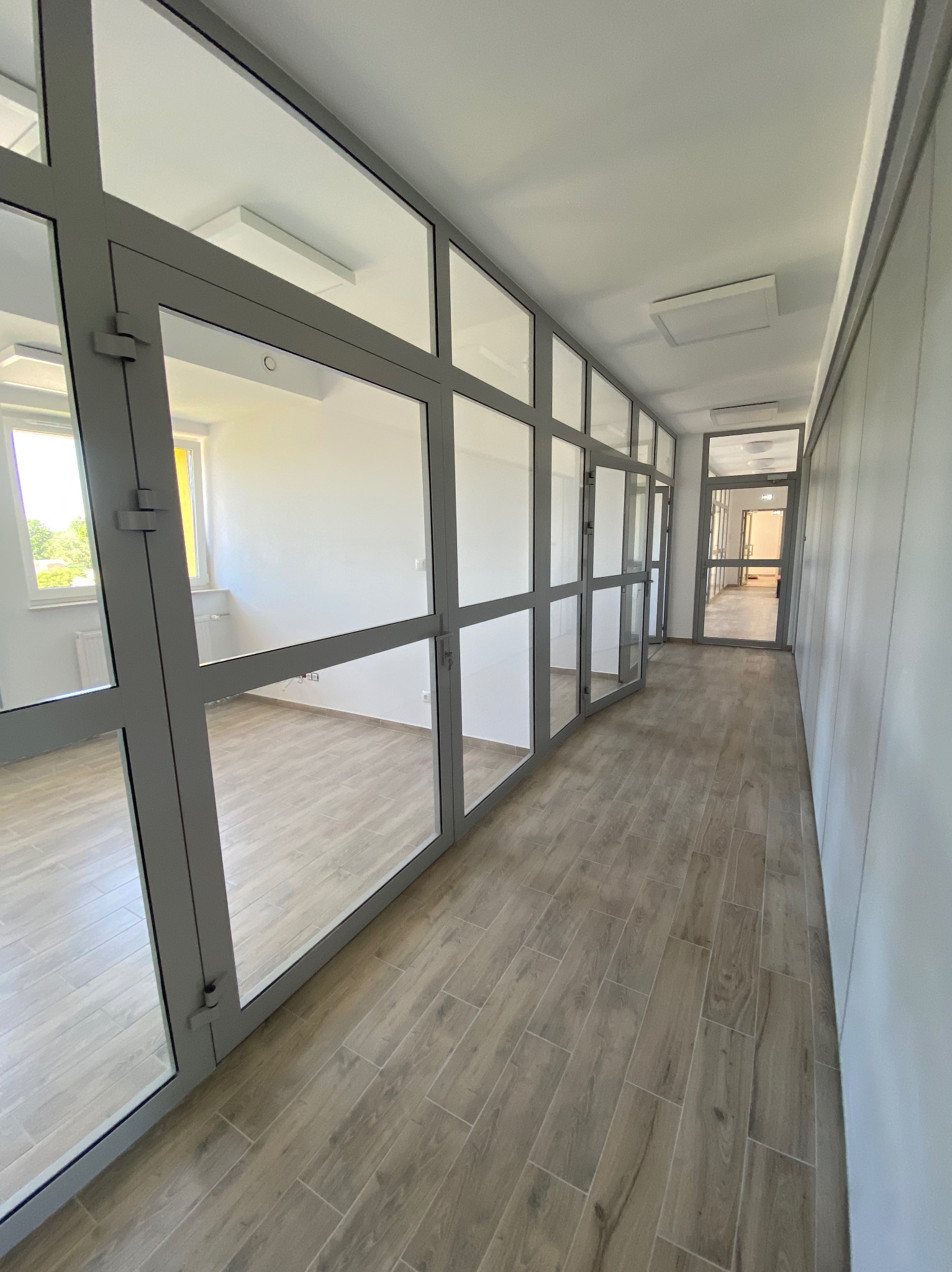 Korytarz z drewnopodobną podłogą, przeszklonymi ścianami działowymi z szarą ramą i białymi ścianami, widok na wnętrza pomieszczeń przez szklane ściany.