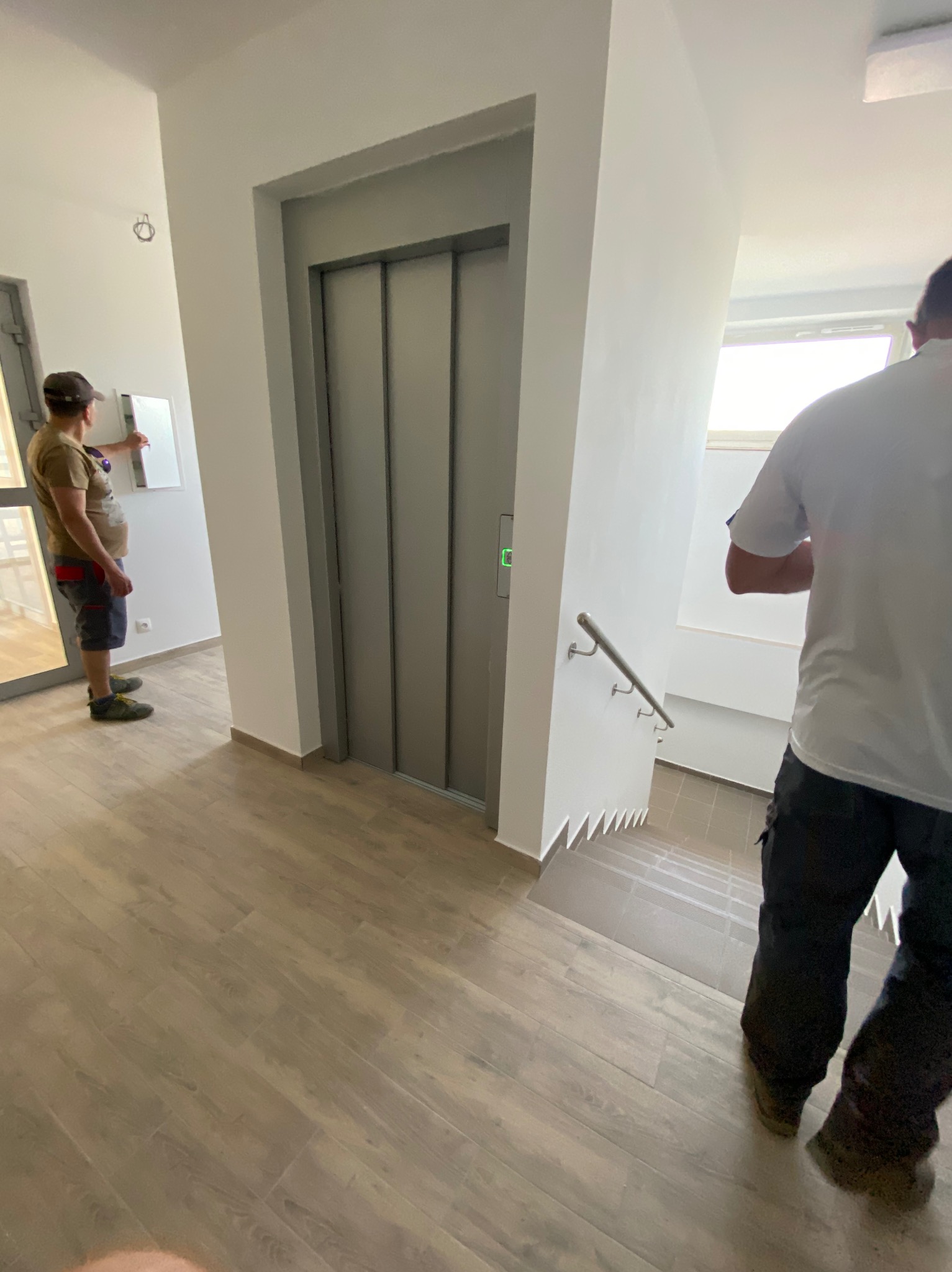 Wnętrze klatki schodowej z windą, pracownik sprawdza skrzynkę elektryczną, widoczne drewnopodobne panele podłogowe i schody wyłożone płytkami.