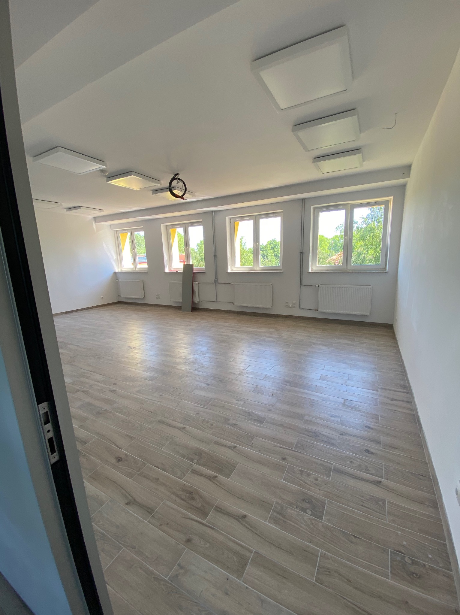 Pusty pokój po remoncie z drewnopodobną podłogą ułożoną w jodełkę, białymi ścianami, grzejnikami pod oknami i nowoczesnym oświetleniem sufitowym.