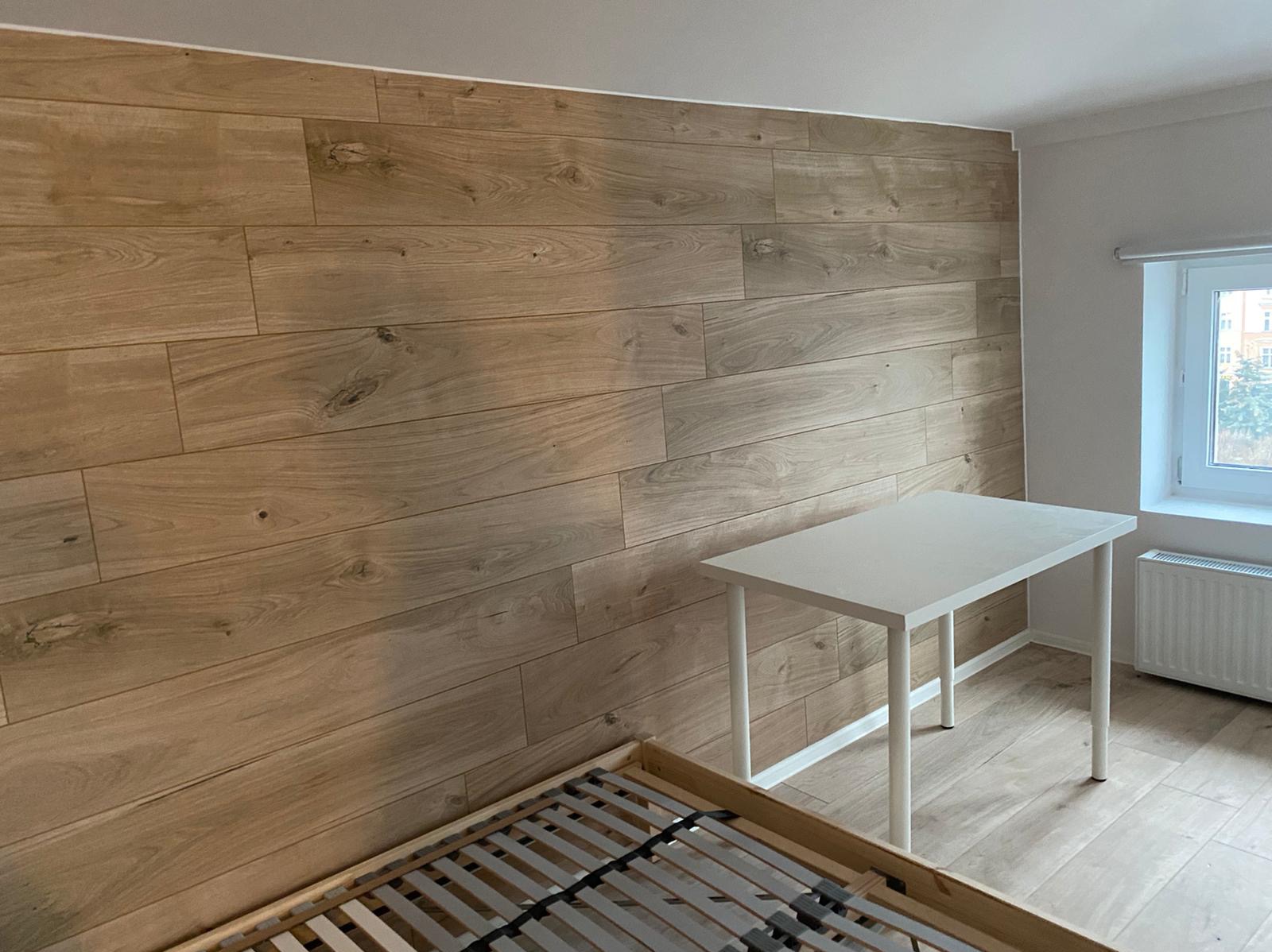 Ściana wykończona panelami drewnopodobnymi ułożonymi poziomo, z widocznym fragmentem łóżka i białym biurkiem w pokoju z oknem.