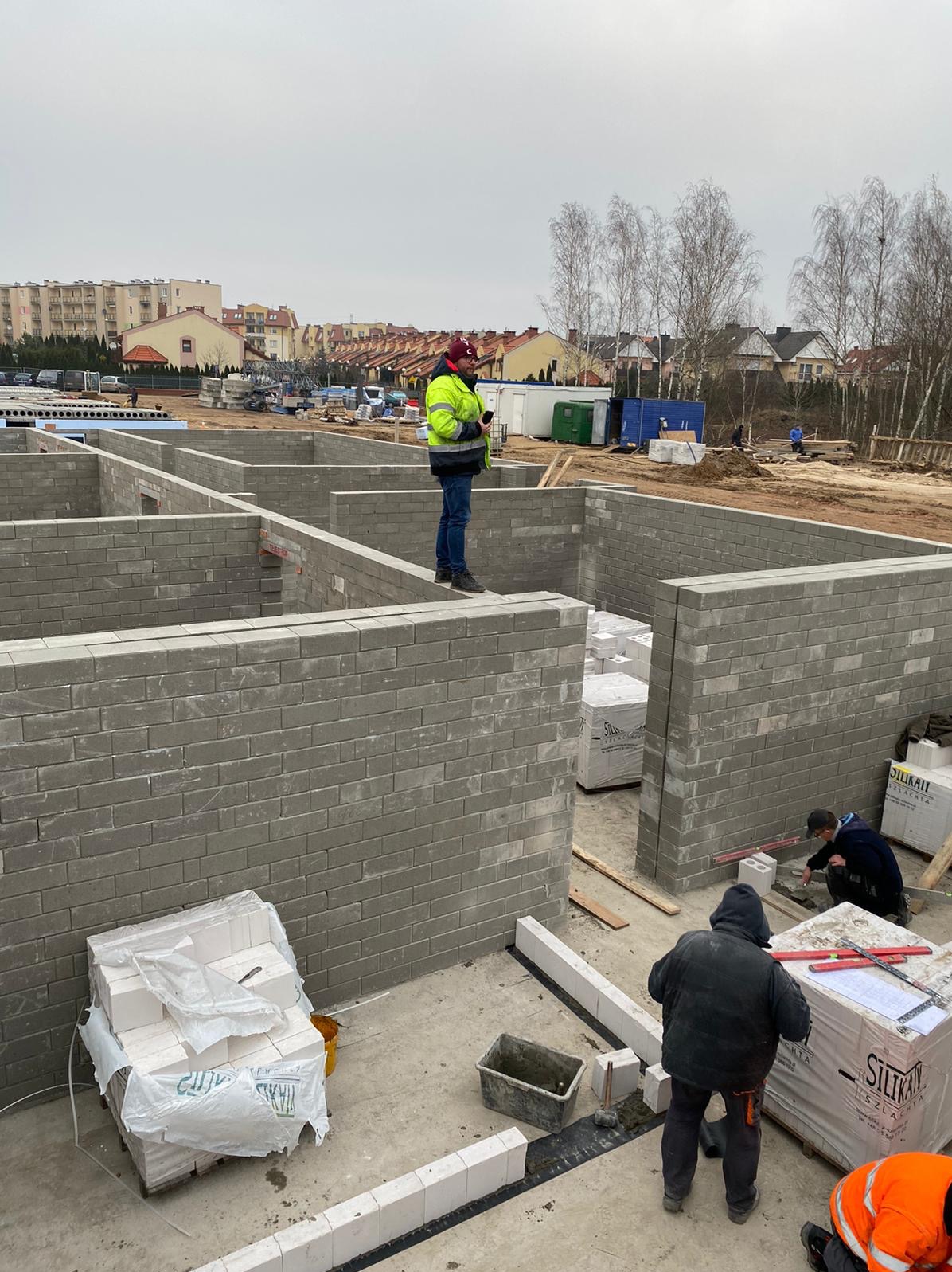 Budowa domu z bloczków silikatowych: widok na fundamenty z murami, pracownicy w odzieży roboczej, palety z bloczkami, otoczenie budowy z budynkami mieszkalnymi w tle.