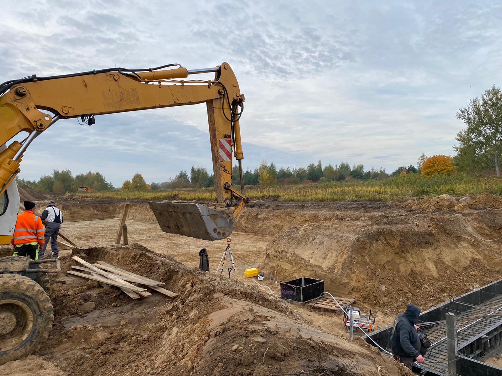Żółta koparka podczas prac ziemnych przy budowie fundamentów domu, widoczni pracownicy w odblaskowych ubraniach, geodezyjny statyw i wykopane rowy.