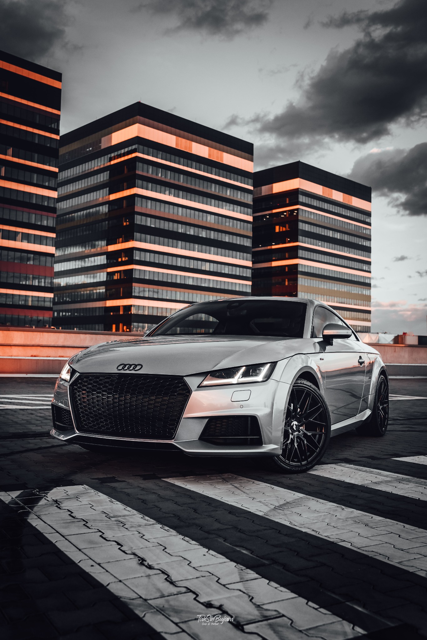 Srebrne Audi TT z czarnymi felgami na tle nowoczesnych budynków biurowych oświetlonych pomarańczowym światłem, zaparkowane na parkingu z pasami dla pieszych.