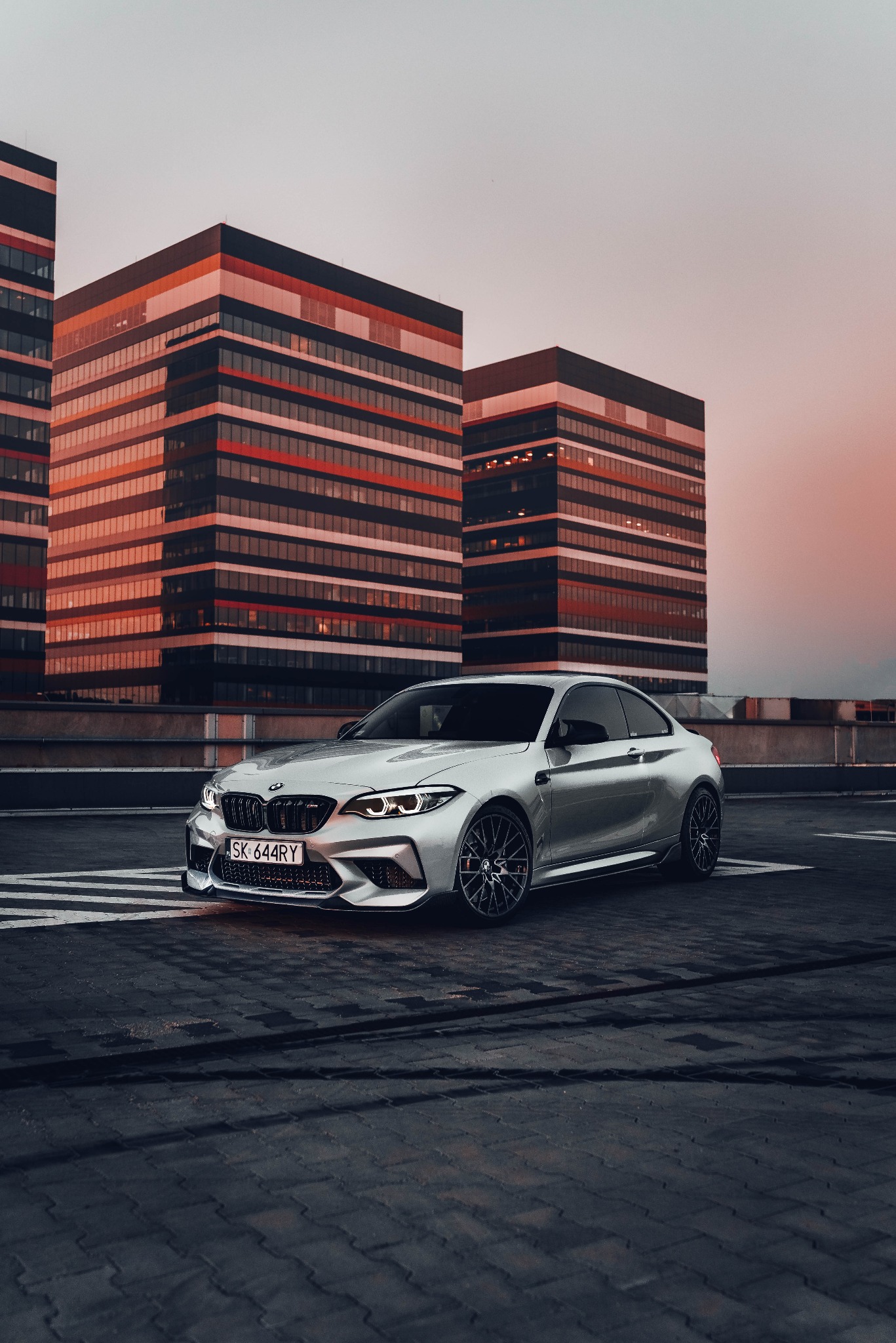 Srebrne sportowe coupe BMW M2 na tle nowoczesnych budynków biurowych o zachodzie słońca.