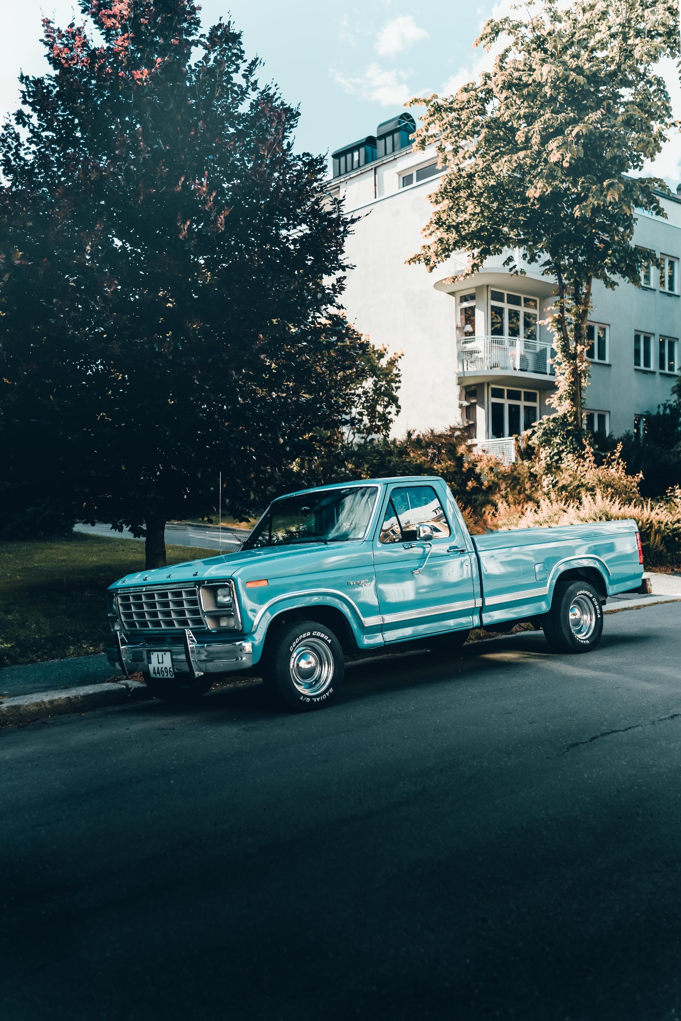 Turkusowy pickup Ford Ranger z lat 80. zaparkowany na ulicy, z budynkiem mieszkalnym i drzewami w tle.
