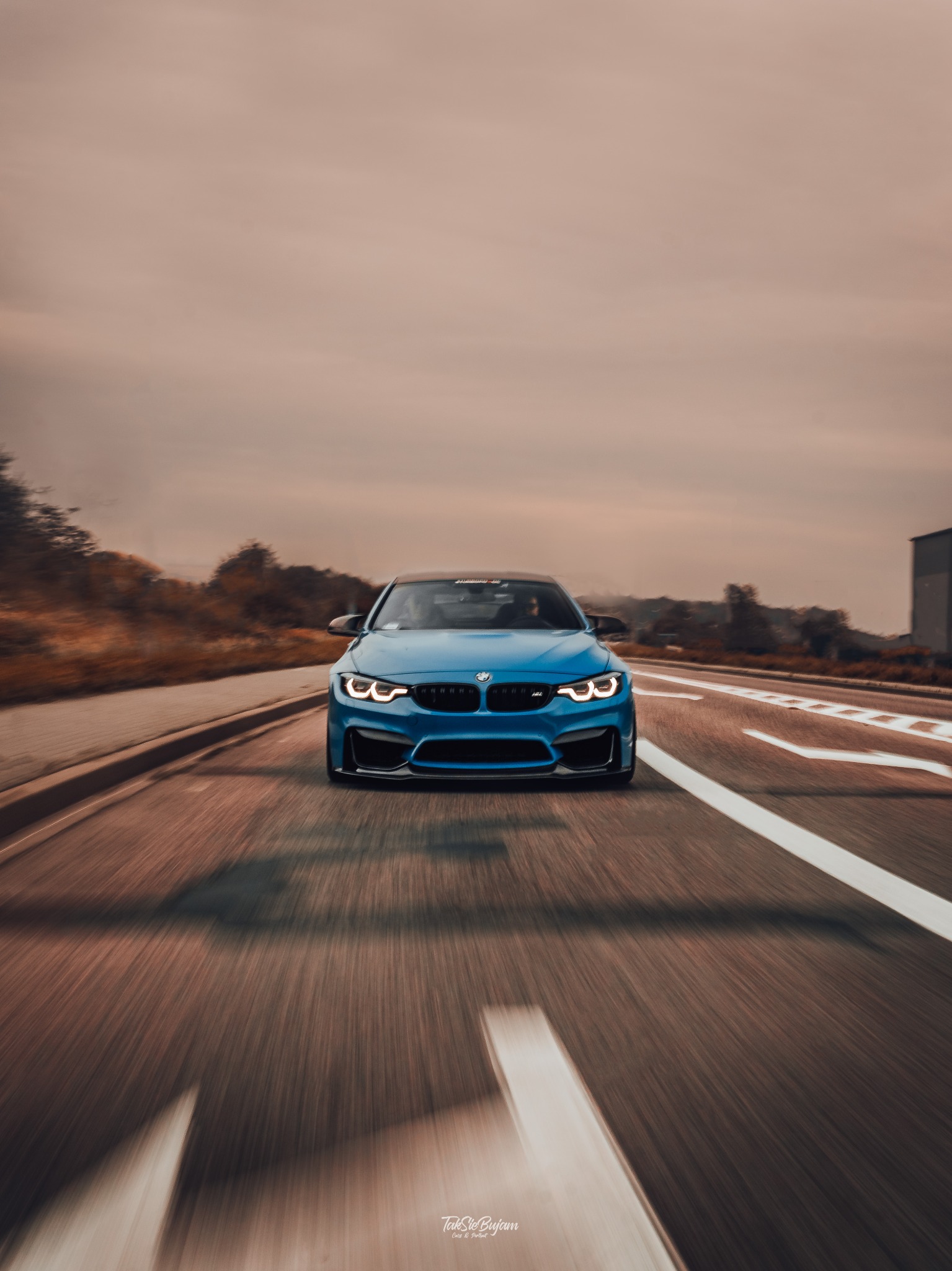 Niebieskie sportowe BMW M3 jedzie po asfaltowej drodze, widok z przodu, dynamiczne rozmycie tła sugeruje dużą prędkość.