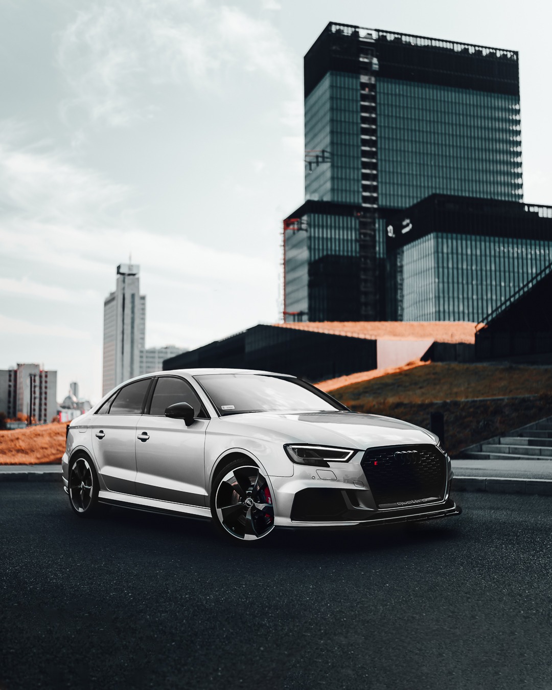 Srebrny samochód Audi RS3 na tle nowoczesnej architektury biurowej, z widocznymi czerwonymi zaciskami hamulcowymi i czarnymi felgami.