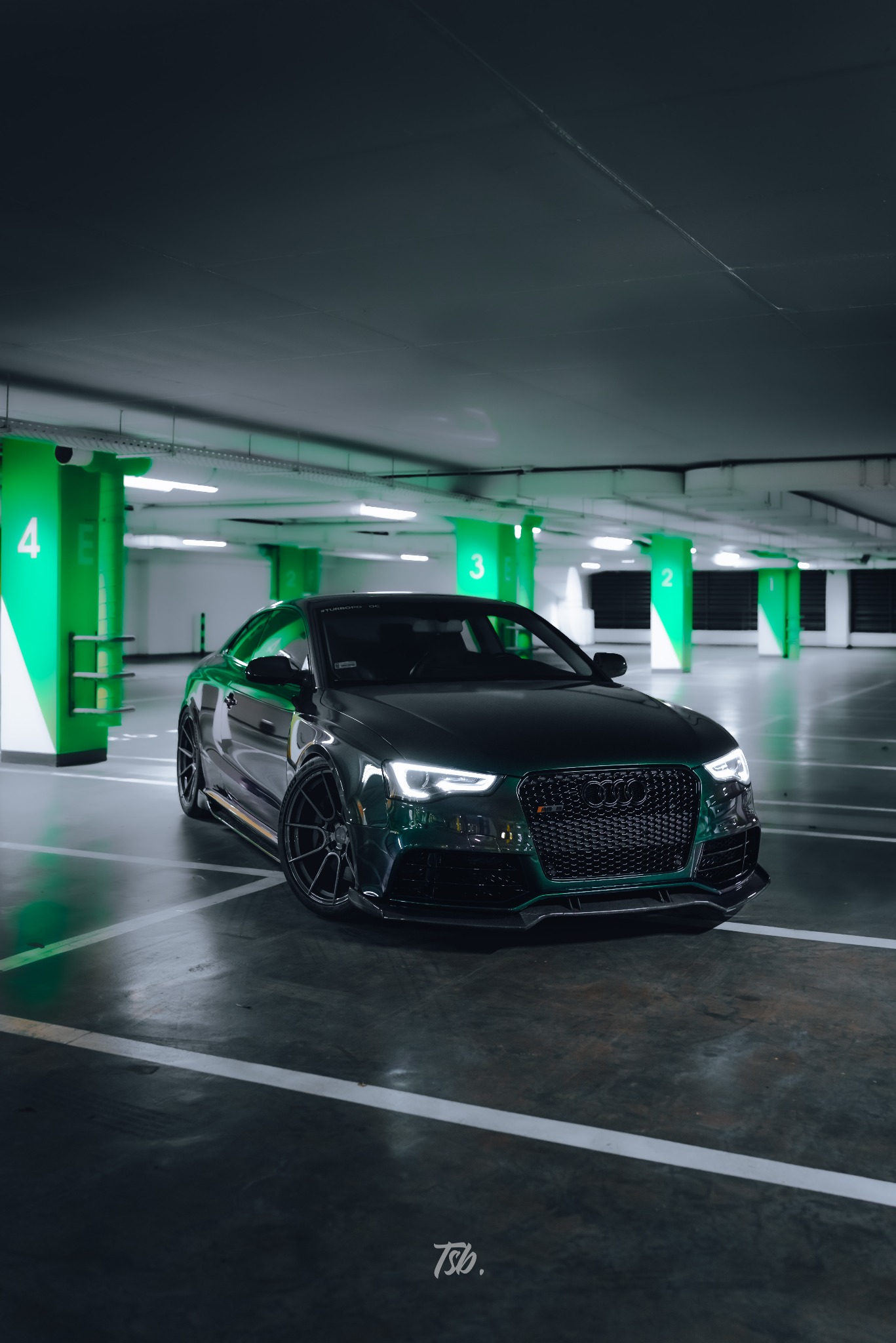 Ciemnozielone Audi RS5 z czarnymi felgami, sfotografowane w garażu podziemnym z zielonym oświetleniem, widok z przodu na zaparkowany samochód.