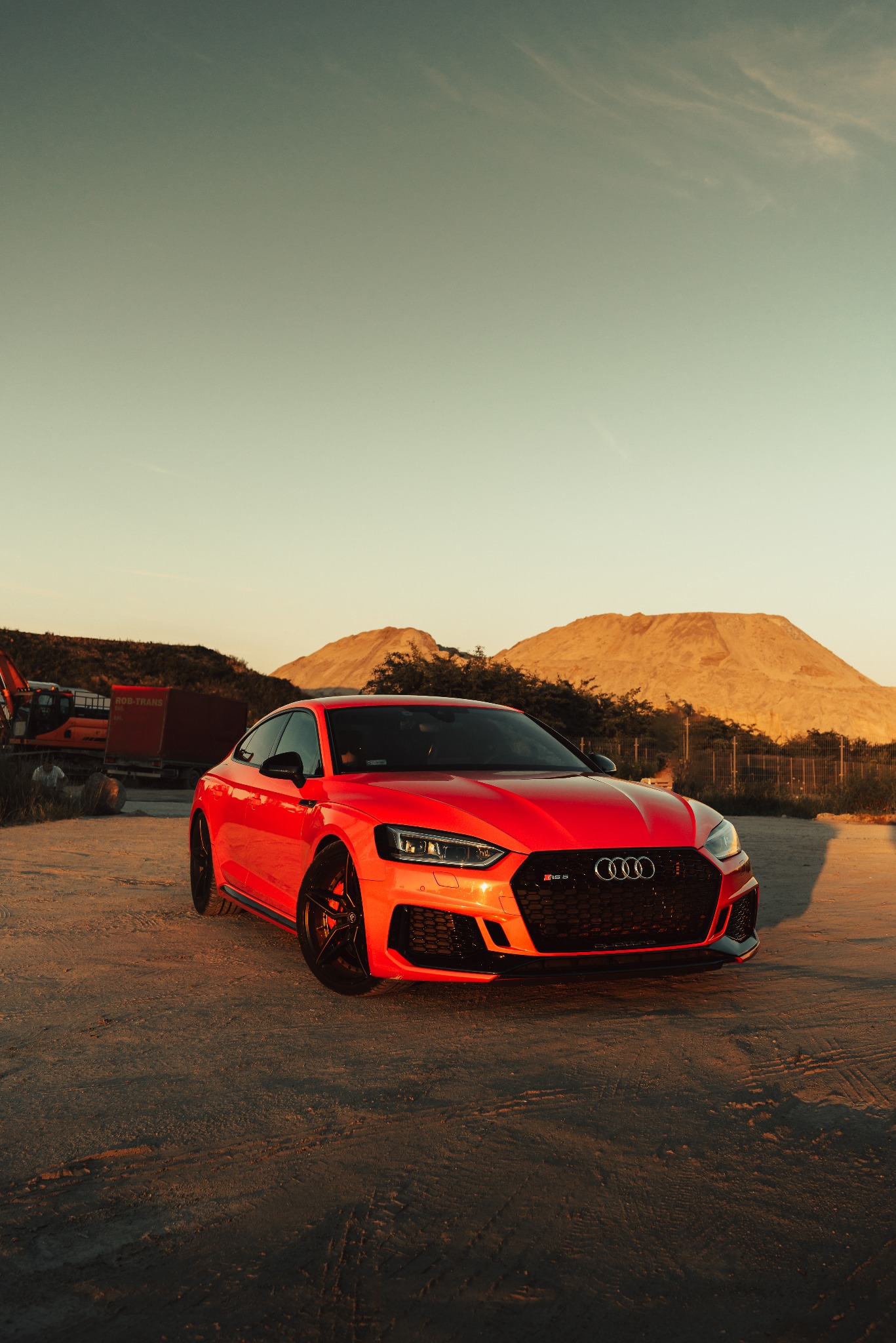 Czerwone Audi RS5 na piaszczystym terenie z kopcami ziemi w tle, sfotografowane w złotym świetle zachodzącego słońca.