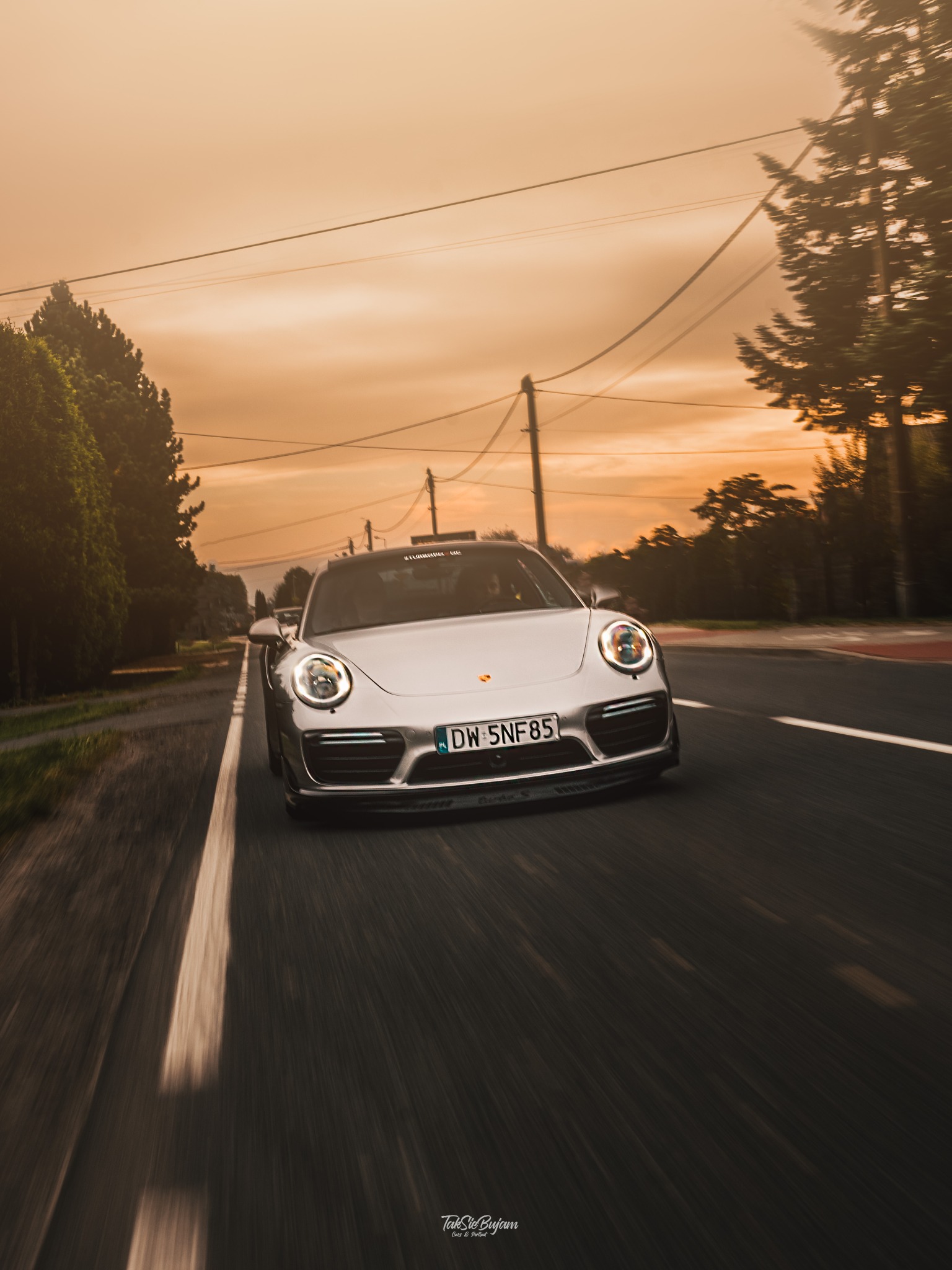 Srebrny samochód sportowy Porsche na drodze, dynamiczne ujęcie z niskiej perspektywy, pomarańczowe niebo o zmierzchu, widoczne drzewa i słupy energetyczne w tle.