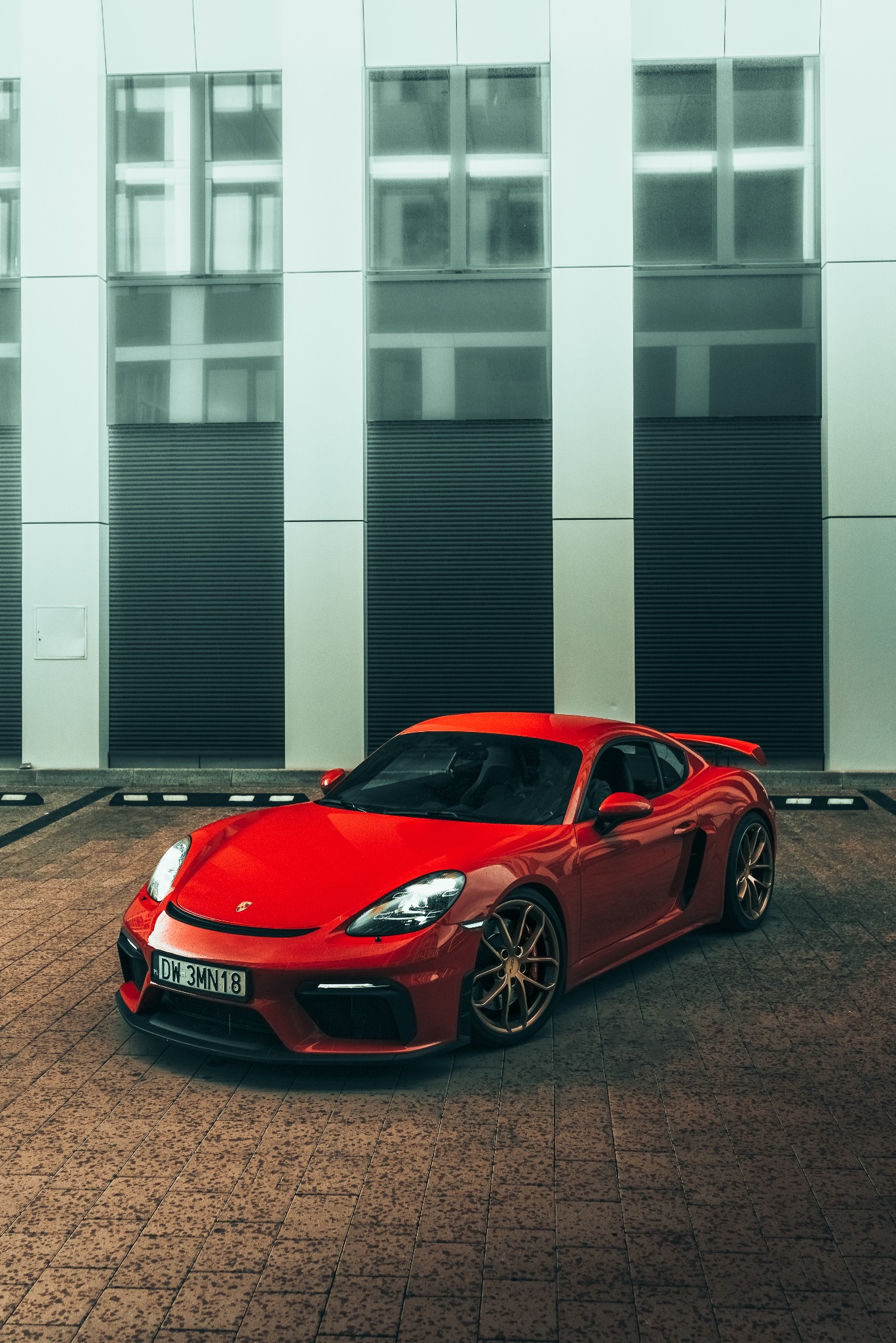 Czerwony sportowy samochód Porsche z brązowymi felgami zaparkowany przed nowoczesnym budynkiem z dużymi oknami.
