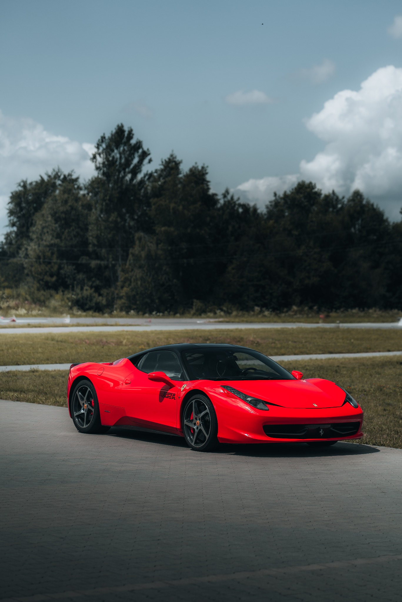 Czerwone Ferrari 458 Italia z czarnym dachem, zaparkowane na szarym asfalcie, z zielonym tłem drzew i pochmurnym niebem.