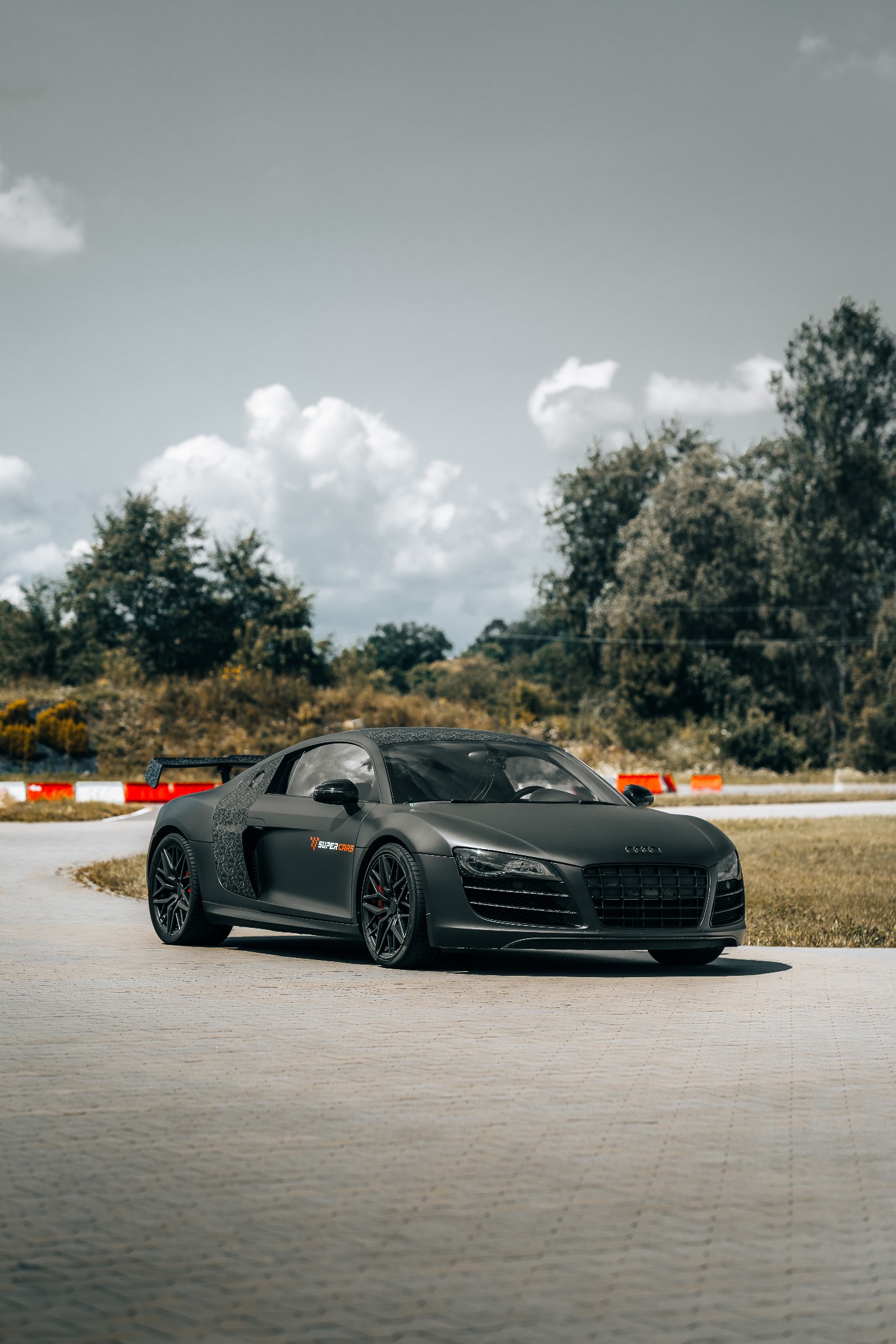 Matowy, czarny Audi R8 z czerwonymi akcentami na felgach, zaparkowany na betonowej nawierzchni toru wyścigowego z widocznymi oponami i trawą w tle.