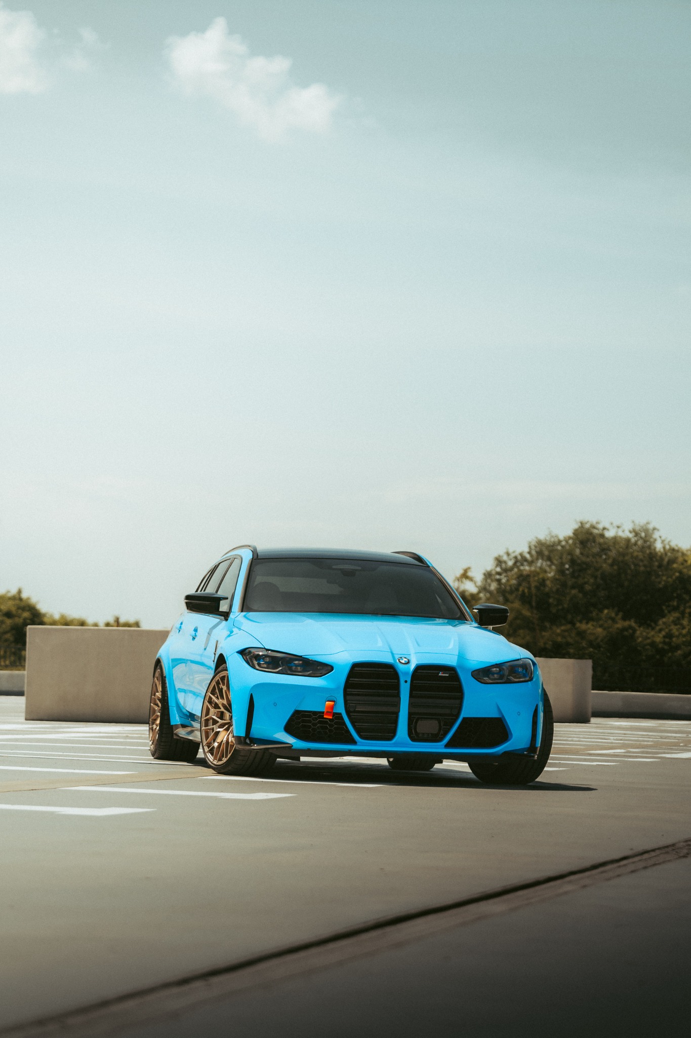 Błękitne sportowe kombi z brązowymi felgami na parkingu, widok z przodu, w tle zieleń drzew.