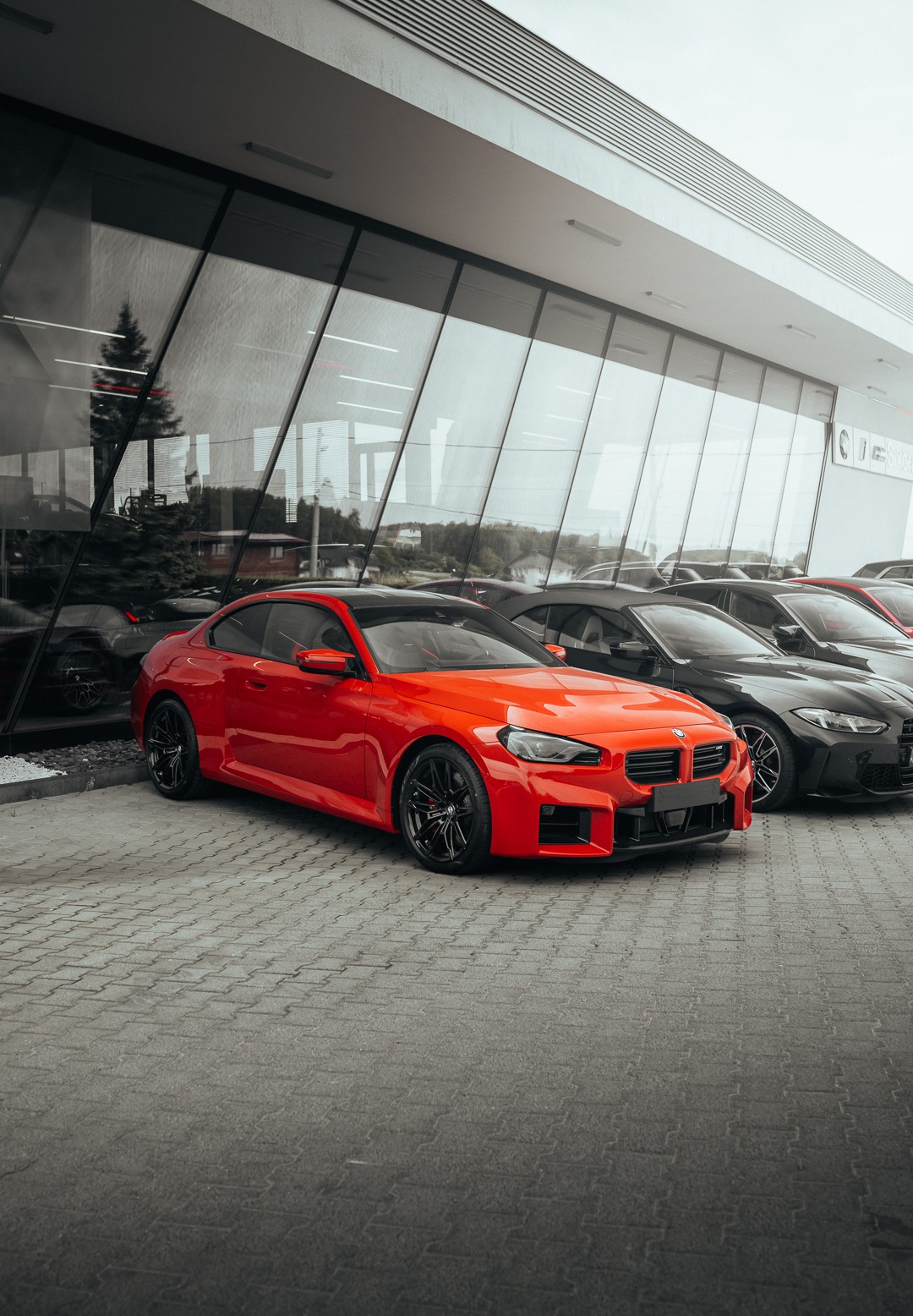 Czerwone sportowe coupe na tle szklanej fasady salonu samochodowego, z widocznymi innymi modelami w tle.