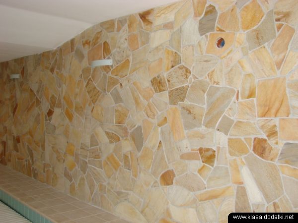 Ściana wyłożona nieregularnymi płytkami kamiennymi w odcieniach beżu i brązu, oświetlona dwoma kinkietami, z widoczną ceramiczną ławką u dołu.