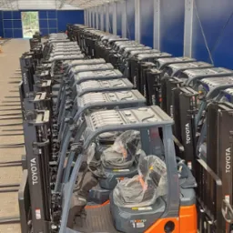 Rząd nowych, pomarańczowo-szarych wózków widłowych Toyota Tonero 15, ustawionych w hali magazynowej z niebieskimi ścianami i białym dachem, fotele owinięte folią ochronną.