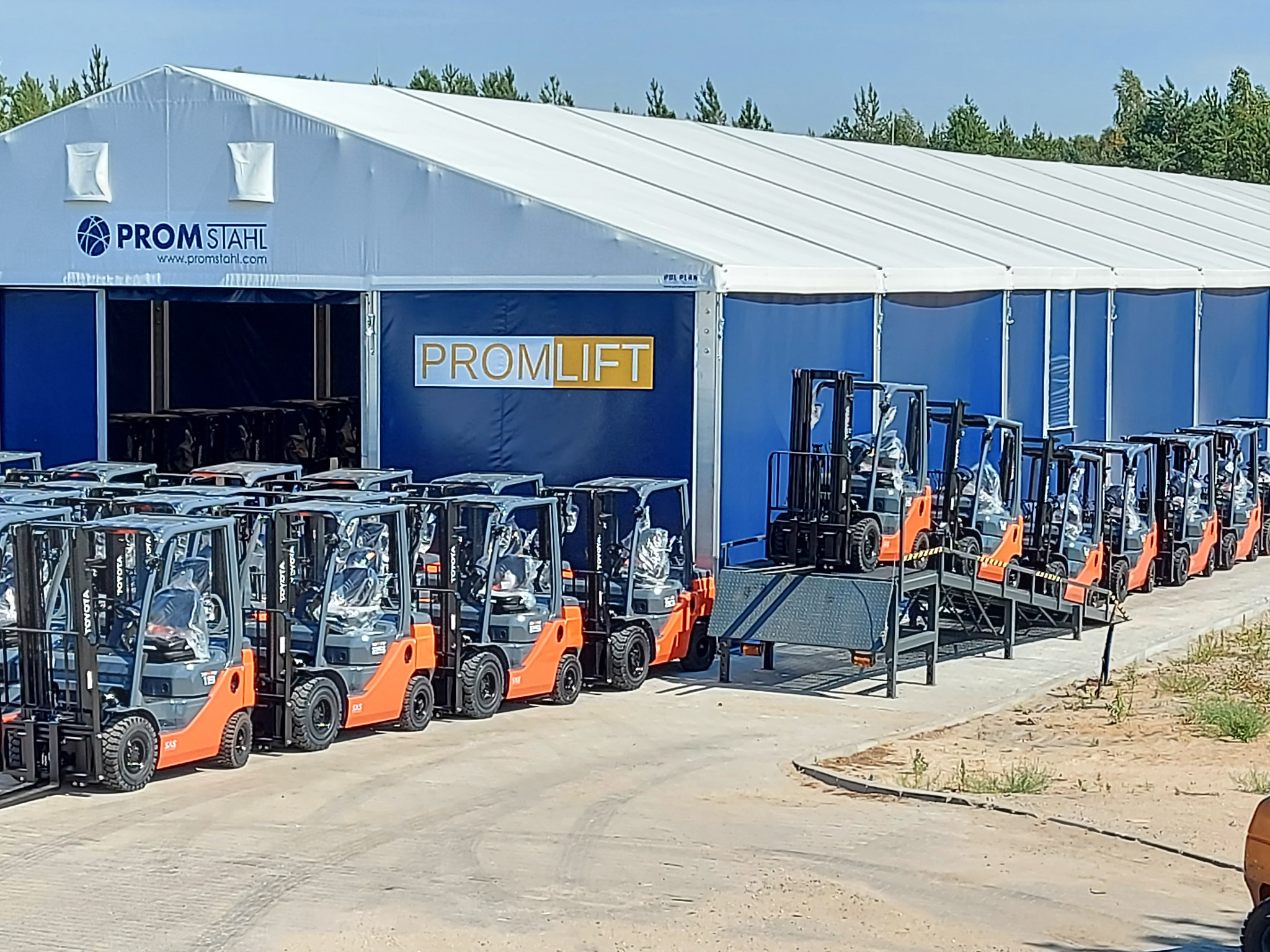 Rząd nowych, pomarańczowych wózków widłowych Toyota, zaparkowanych przed magazynem z logo 'PROMLIFT' i 'PROM-STAHL', gotowych do dystrybucji.