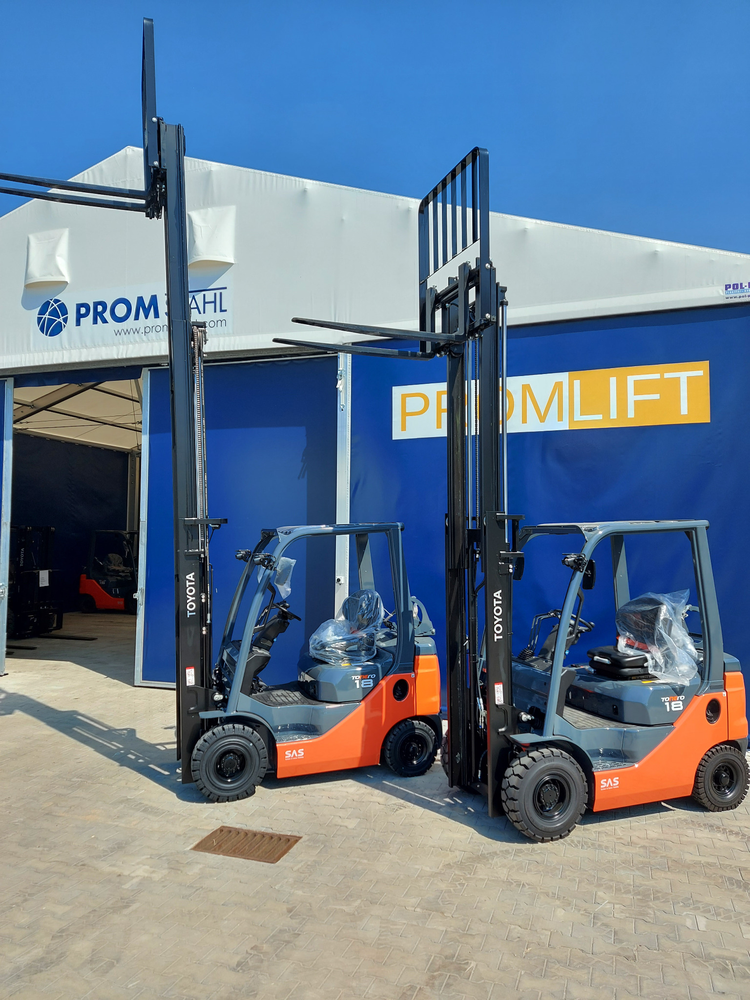 Dwa nowe wózki widłowe Toyota stojące przed halą z logo Promlift, jeden z wózków ma jeszcze folię ochronną na siedzeniu.