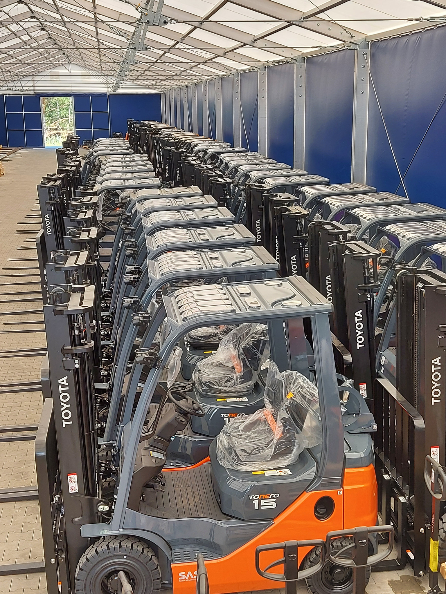 Rząd nowych, pomarańczowo-szarych wózków widłowych Toyota Tonero 15, ustawionych w hali magazynowej z niebieskimi ścianami i białym dachem, fotele owinięte folią ochronną.