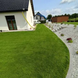 Widok na zadbany trawnik przy domu z białymi ścianami i ciemnym dachem. Granica trawnika wyłożona szarymi kamieniami, a w tle widać rośliny i budynek w budowie.