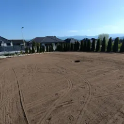 Przygotowany teren pod trawnik: wyrównana ziemia z widocznymi śladami maszyn, rząd posadzonych drzew iglastych wzdłuż ogrodzenia, błękitne niebo.