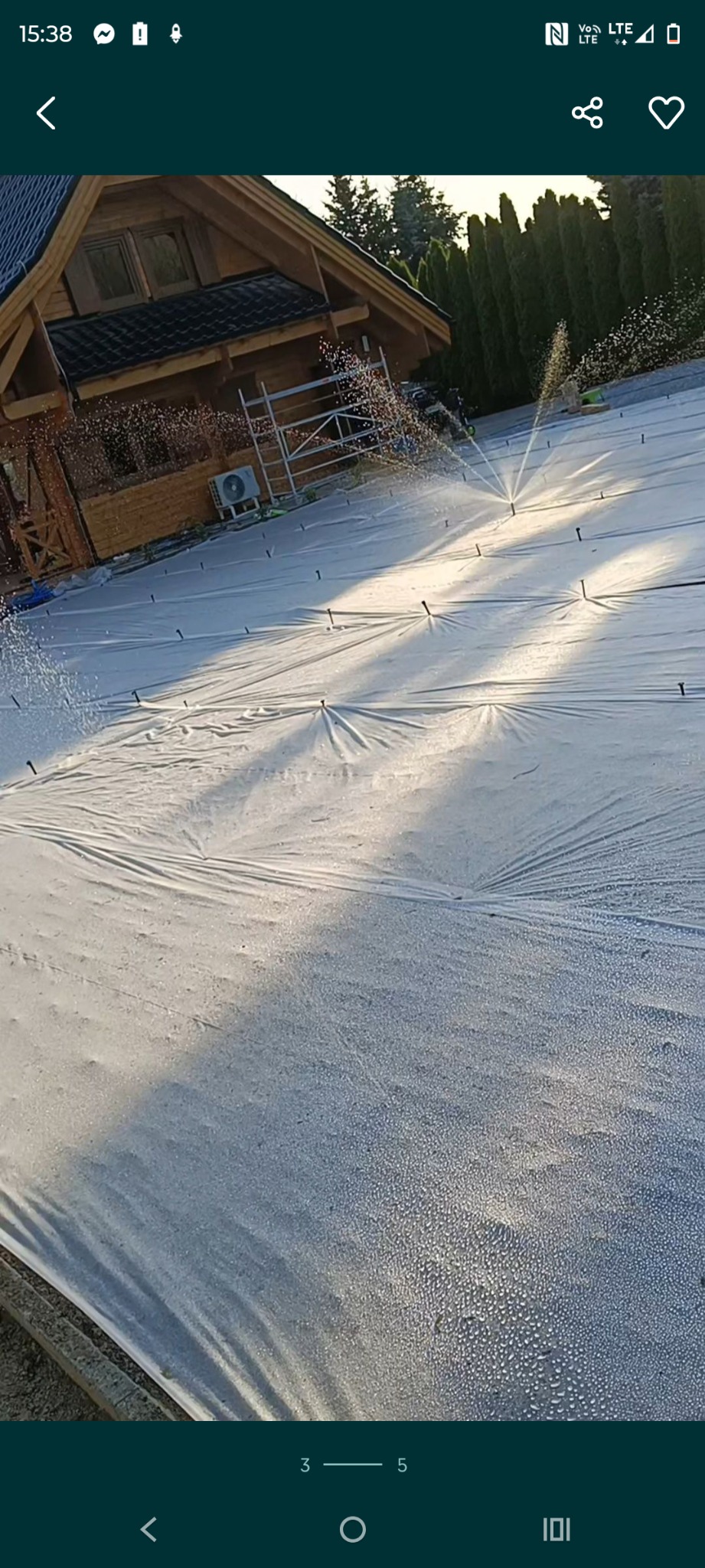 Instalacja nawadniania pod trawnik z rolki. System zraszający w trakcie pracy, teren pokryty folią, drewniany dom w tle, wysokie tuje po prawej.