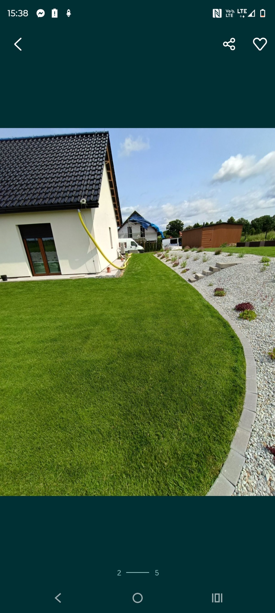 Widok na zadbany trawnik przy domu z białymi ścianami i ciemnym dachem. Granica trawnika wyłożona szarymi kamieniami, a w tle widać rośliny i budynek w budowie.