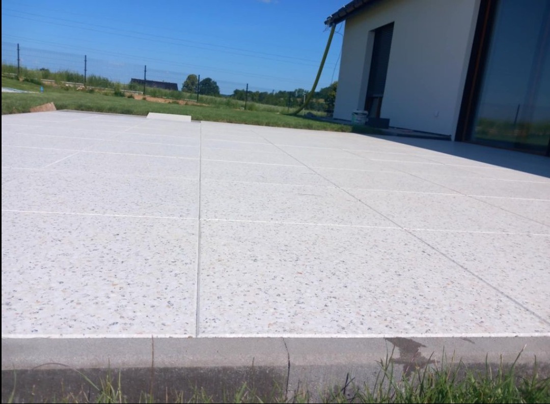 Wykończony taras z jasnych, kwadratowych płyt z drobnym wzorem, położonych na równym podłożu. Widoczny fragment budynku z dużym oknem i trawnik w tle.