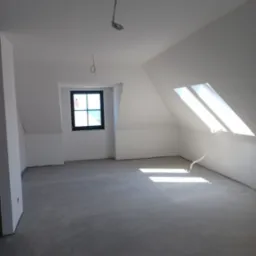 Poddasze po wykonaniu prac wykończeniowych, ściany i sufit pomalowane na biało, szary betonowy podkład, widoczne okno dachowe i zwykłe okno w czarnej ramie, zwisające kable oświetleniowe.