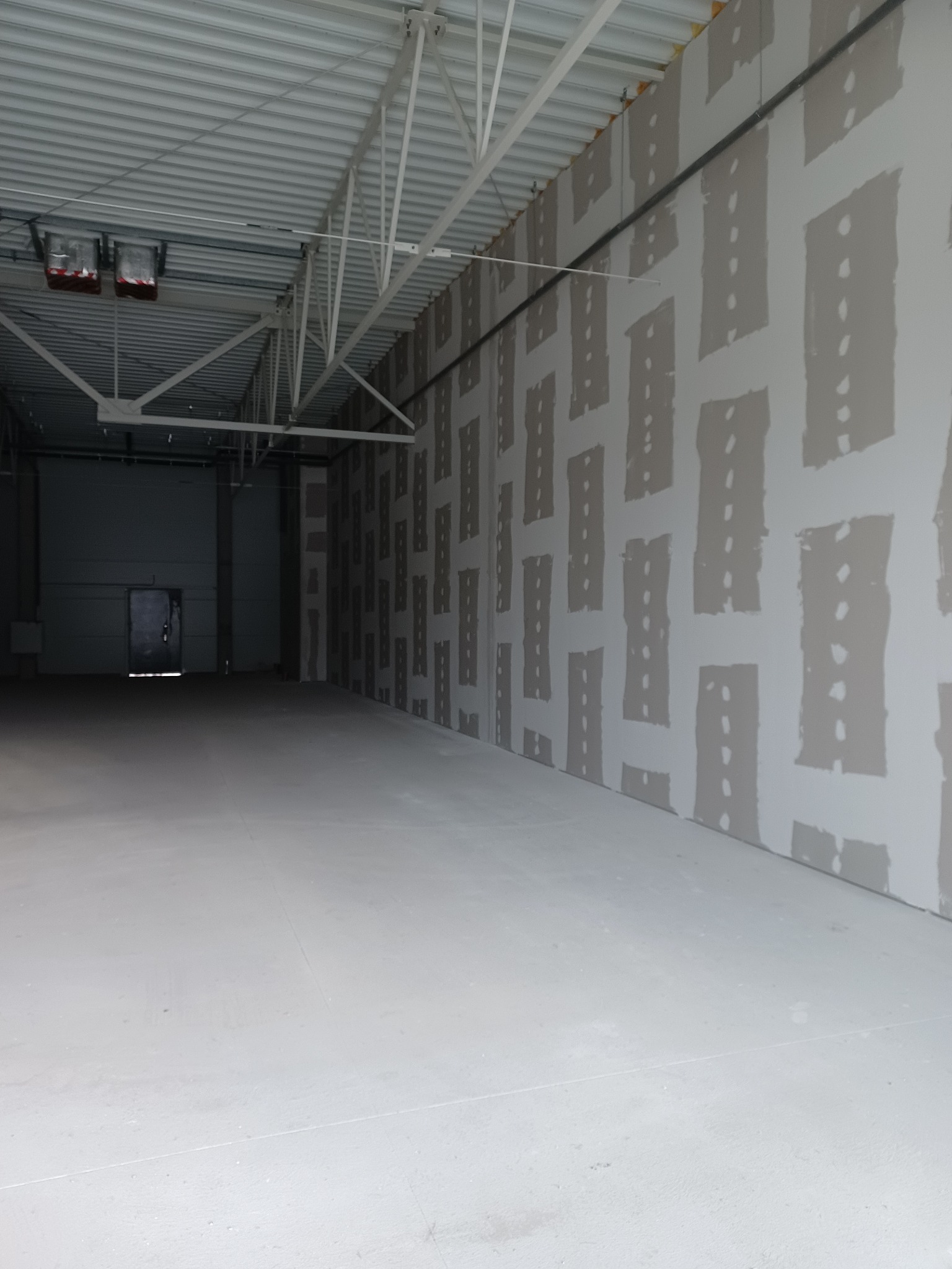 Wnętrze hali produkcyjnej z zamontowanymi płytami na ścianach, częściowo pokrytymi masą wyrównującą, widoczne szpary między płytami wypełnione masą.