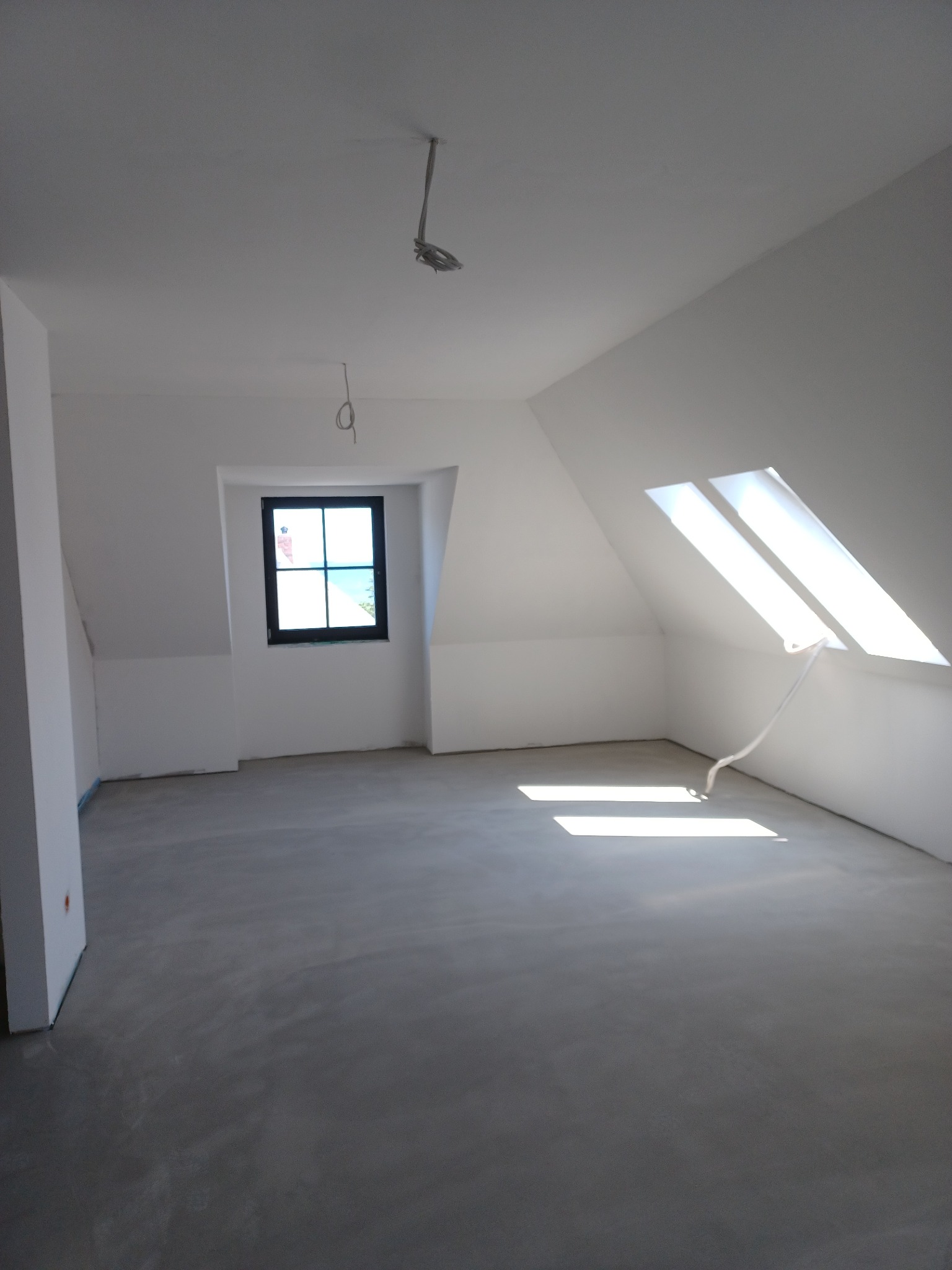 Poddasze po wykonaniu prac wykończeniowych, ściany i sufit pomalowane na biało, szary betonowy podkład, widoczne okno dachowe i zwykłe okno w czarnej ramie, zwisające kable oświetleniowe.