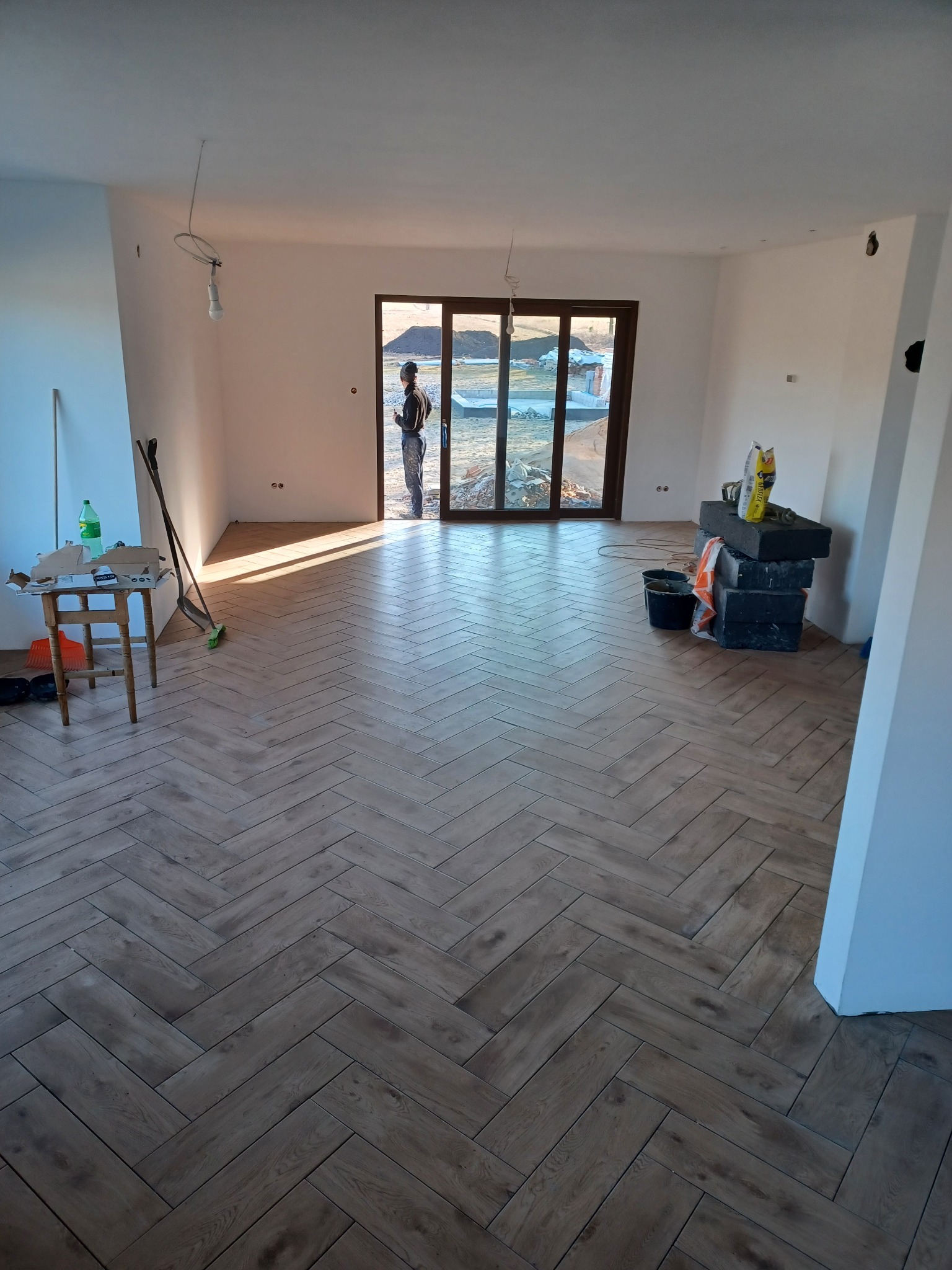 Wnętrze w trakcie remontu z ułożoną podłogą w jodełkę, widoczne narzędzia, worki z zaprawą i osoba stojąca w drzwiach balkonowych.