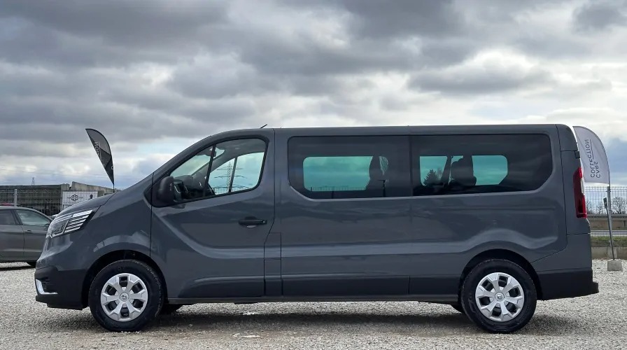 Szary bus z przyciemnianymi szybami, widok z boku na tle pochmurnego nieba i żwirowego podłoża, z widocznym banerem reklamowym w tle.