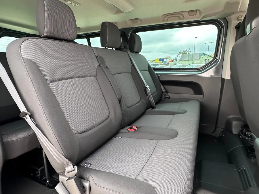 Wnętrze szarego busa z trzema rzędami siedzeń, widok na tylne siedzenia z pasami bezpieczeństwa, przez okno widoczny parking.