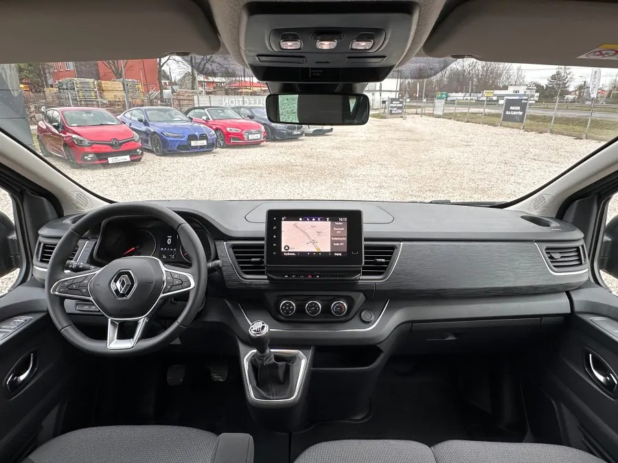 Wnętrze Renault Trafic z manualną skrzynią biegów, widok na deskę rozdzielczą i system nawigacji, z zewnątrz widoczne zaparkowane samochody osobowe.