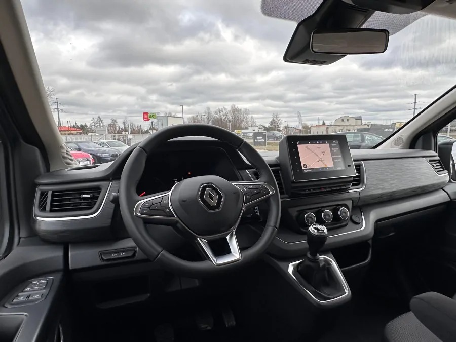 Wnętrze Renault Trafic z manualną skrzynią biegów, widok na deskę rozdzielczą i system nawigacji, na zewnątrz pochmurne niebo.