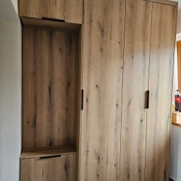 Meble JM - Michał Wyka - Drewniana szafa z czarnymi uchwytami, z otwartą półką i szufladą po lewej stronie. Widoczne butelki przy oknie. Ujęcie z dołu, podkreślające wysokość mebla.