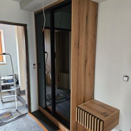 Meble JM - Michał Wyka - Montaż szafy przesuwnej z drewnianym dekorem i czarnymi ramami. Widoczna szafka nocna z lamelami. W tle drabina i fragment remontowanego pomieszczenia.