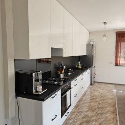 Meble JM - Michał Wyka - Biała kuchnia z połyskiem, czarny blat, nowoczesne uchwyty. Ekspres do kawy na blacie. Widok na okno z drewnianymi żaluzjami. Minimalistyczny design.
