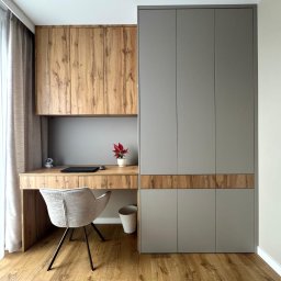 Meble JM - Michał Wyka - Nowoczesny pokój z biurkiem z drewnianym blatem i szarą szafą w zabudowie. Na biurku laptop, roślina w doniczce i figurka. Jasna podłoga i krzesło.