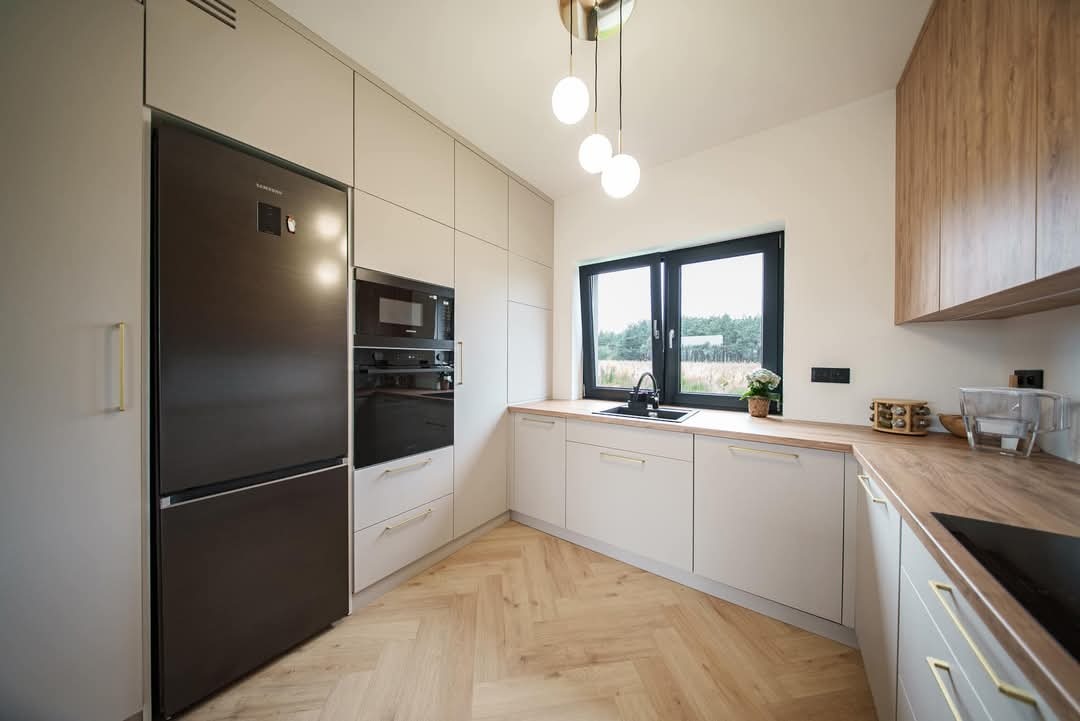 Jasna kuchnia z drewnianym blatem i podłogą w jodełkę. Czarna armatura i okno. Minimalistyczne szafki z długimi, cienkimi uchwytami. Nowoczesne oświetlenie.