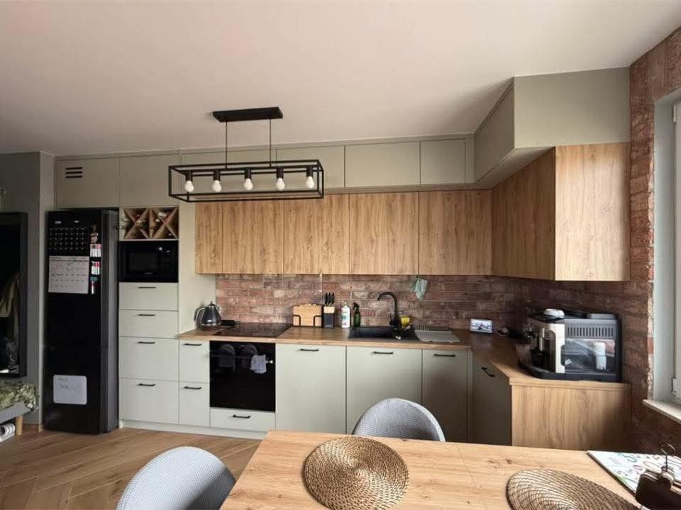 Nowoczesna kuchnia z frontami w odcieniach zieleni i drewna, ceglana ściana nad blatem, czarny sprzęt AGD i lampa. Widok na stół z plecionymi podkładkami.