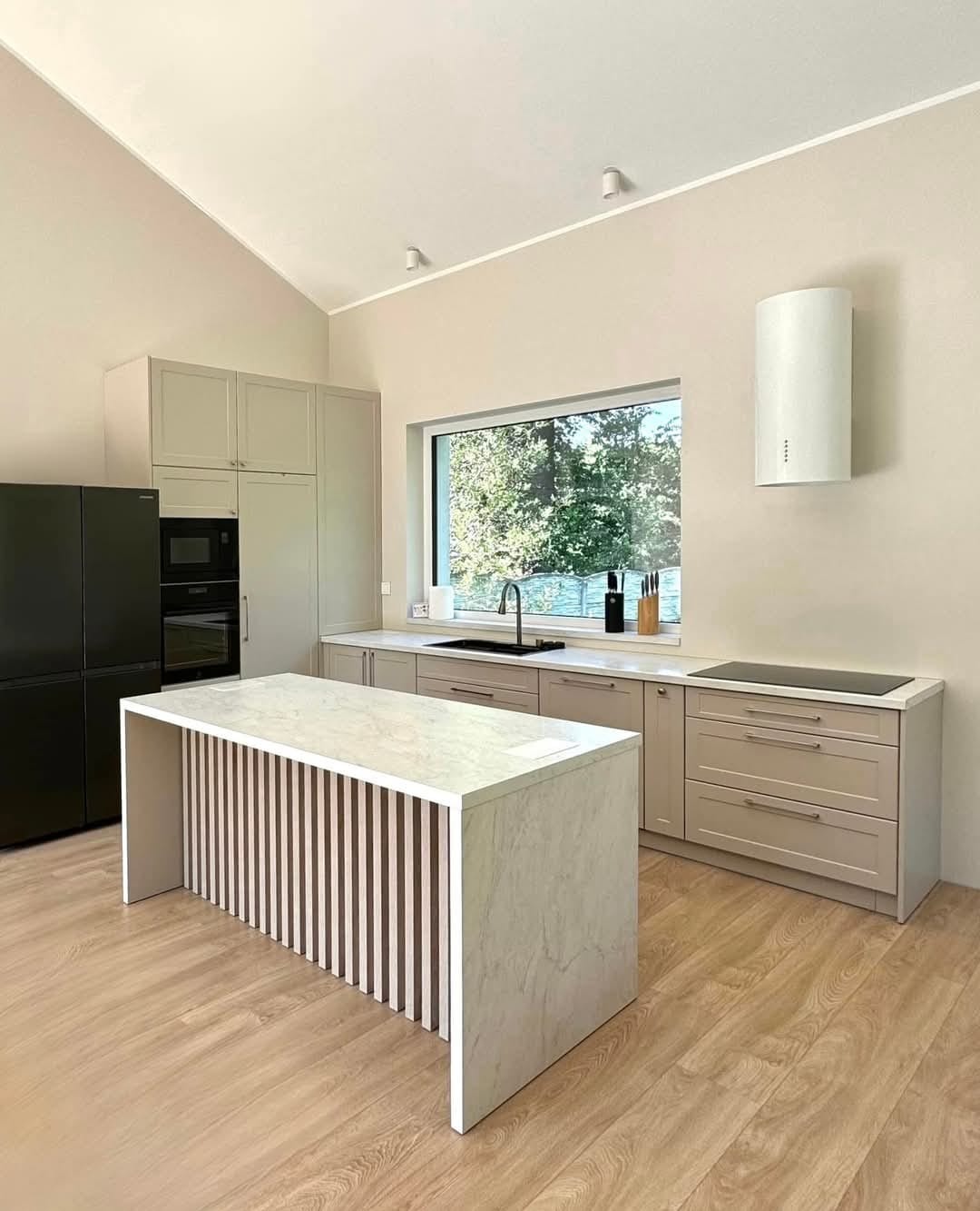Jasna kuchnia z wyspą o marmurowym blacie i pionowymi listwami. Minimalistyczny design, okno z widokiem na zieleń. Nowoczesny okap cylindryczny.