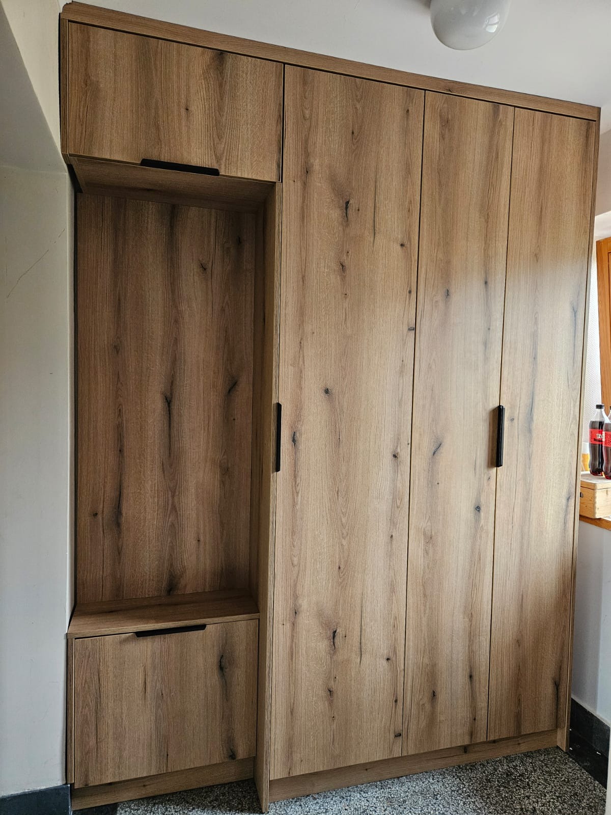 Drewniana szafa z czarnymi uchwytami, z otwartą półką i szufladą po lewej stronie. Widoczne butelki przy oknie. Ujęcie z dołu, podkreślające wysokość mebla.